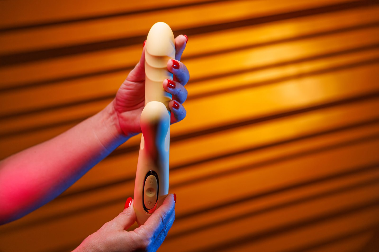 A woman with sexy red nails displays a yellow vibrator turning it in her hands. Neon lighting adds emphasis to the vibrator and hands against a bright yellow lit backdrop.