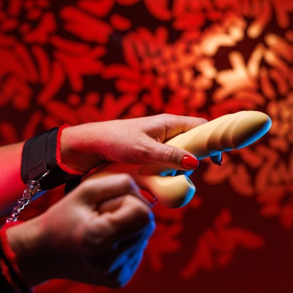A womans hands in soft handcuffs showcase her bold red manicure while holding a yellow clitoral vibrator The dreamy red lit backdrop sets the stage for intimate exploration
